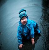 Child sitting in puddle wearing Muddy Buddy waterproof coveralls in blue